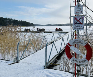Snöklädd brygga som leder till sjö där isen ligger. I förgrunden livboj på ställning.  