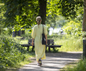 Sommarklädd kvinna vandrar på stig genom grönt landskap. 