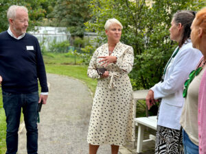 Äldre- och socialförsäkringsminister Anna Tenje i vit klänning samspråkar med Andreas Frykstrand, Helena Gille och Monica Lyander på gården utanför äldreboendet Villa Skönviken.