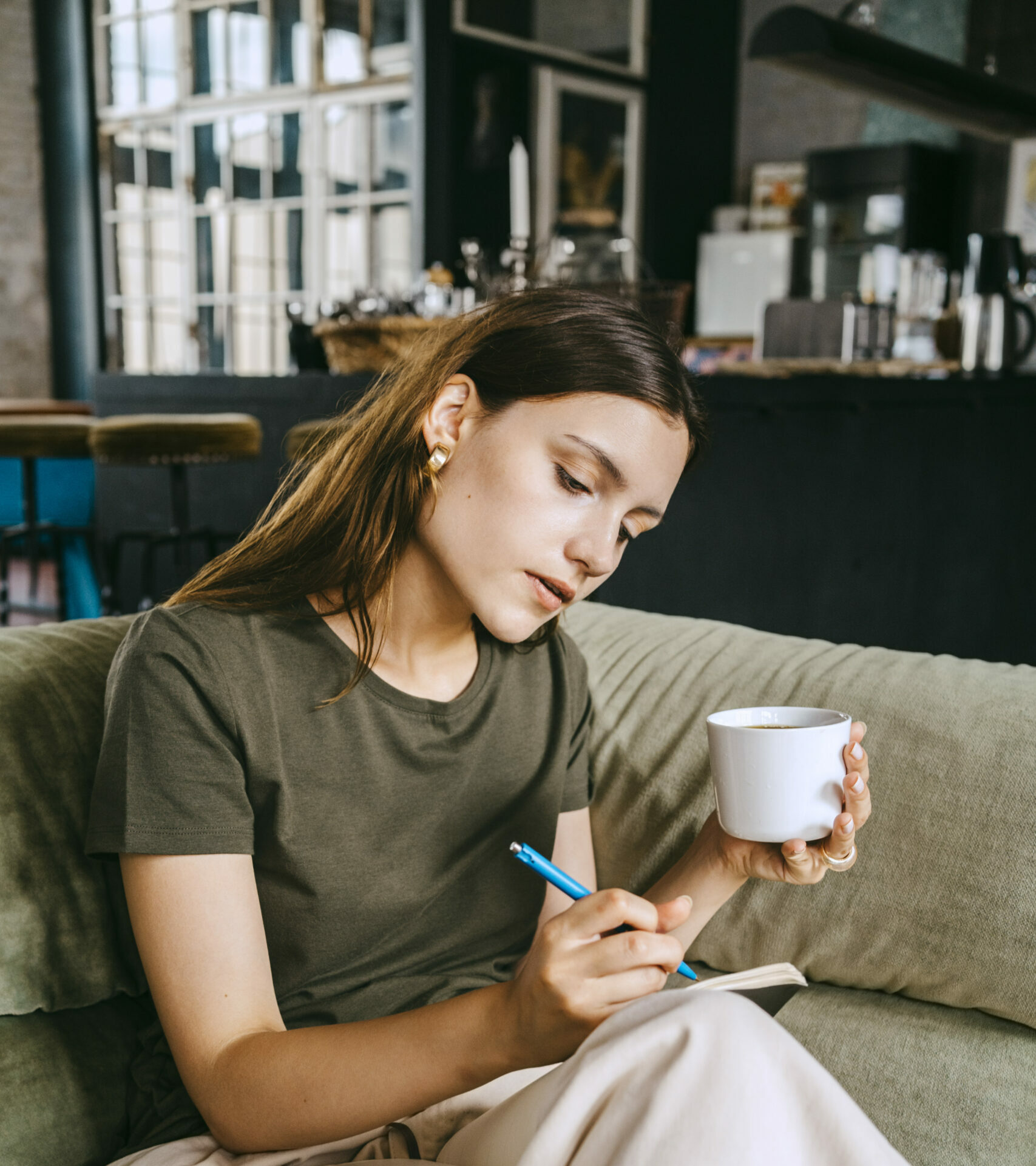 Ung kvinna skriver i en bok med en kaffemugg i handen medan hon sitter på en soffa.