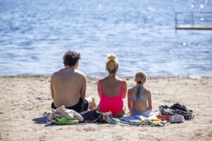 Tre personer i badkläder sitter på sandstrand och kikar ut över sjön Drevviken.